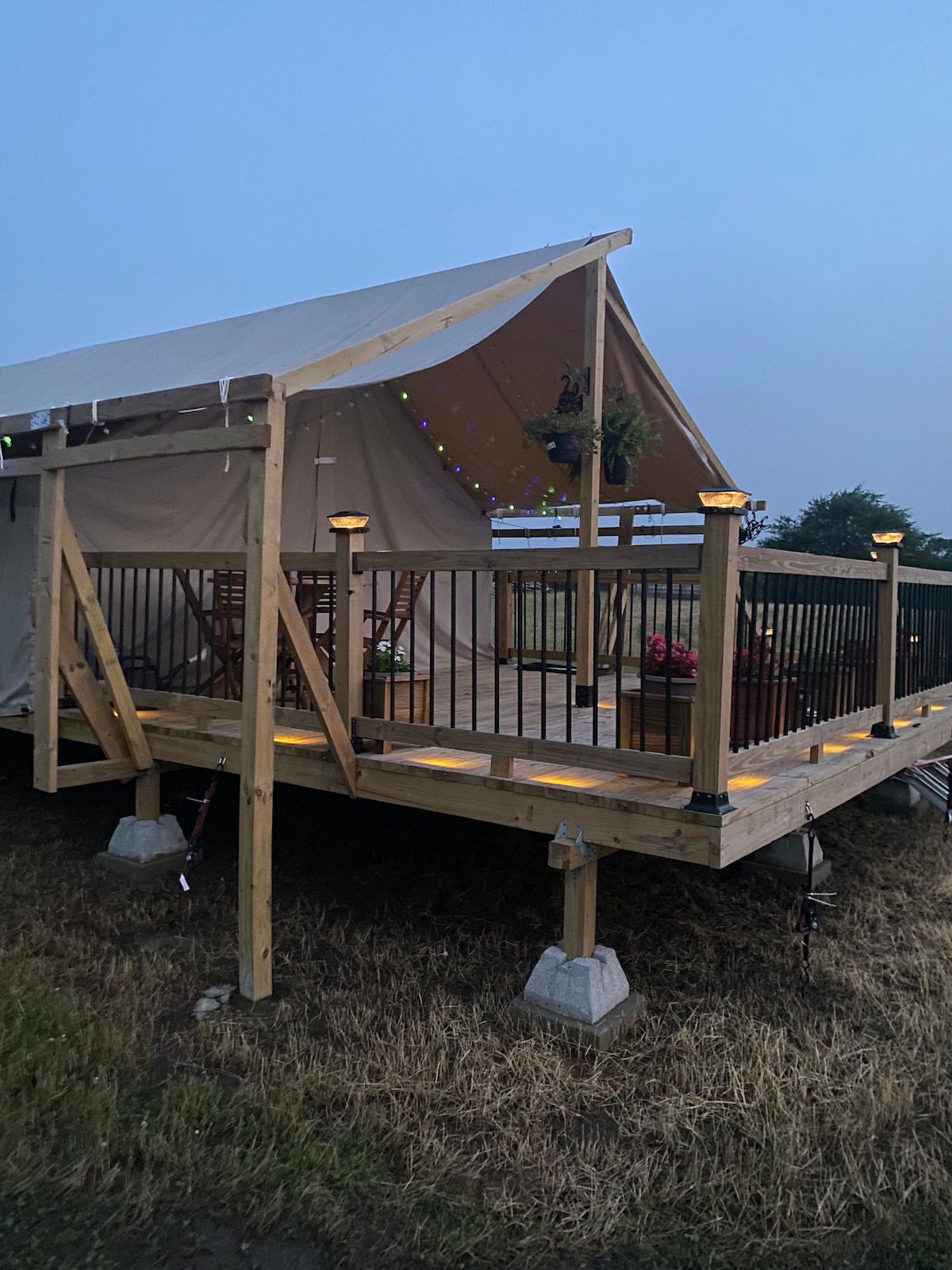 A spacious tent is situated on a wooden deck, featuring a railing adorned with potted plants. Soft lighting along the deck creates a welcoming ambiance. A small table with chairs is visible for outdoor dining, surrounded by natural grass and open space.