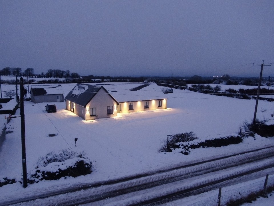 A spacious house is surrounded by a blanket of snow, illuminated by soft lights along its exterior. The serene landscape includes snow-covered fields and a pathway lined with power poles, providing a tranquil winter ambiance.