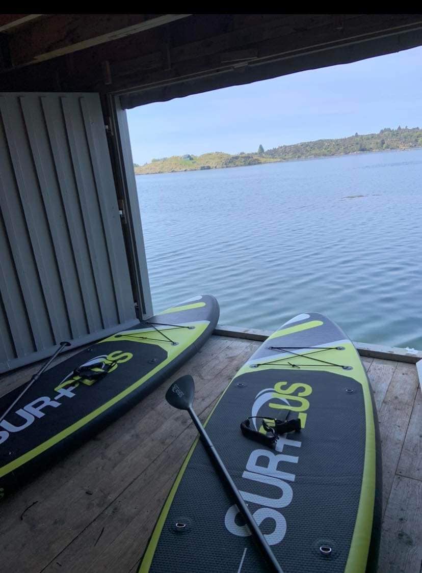 Two paddleboards are positioned inside a boathouse, facing an expansive view of calm waters. Natural light filters through an open window, creating a serene atmosphere. A paddle rests beside the boards on the wooden floor, contributing to the waterfront setting.
