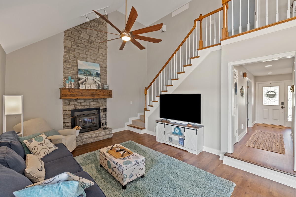 A spacious living area features a stone fireplace with a wooden mantel, complemented by an overhead ceiling fan. A comfortable sofa is positioned on the soft rug, and a large television is mounted on a media console. Natural light flows in from the entrance.