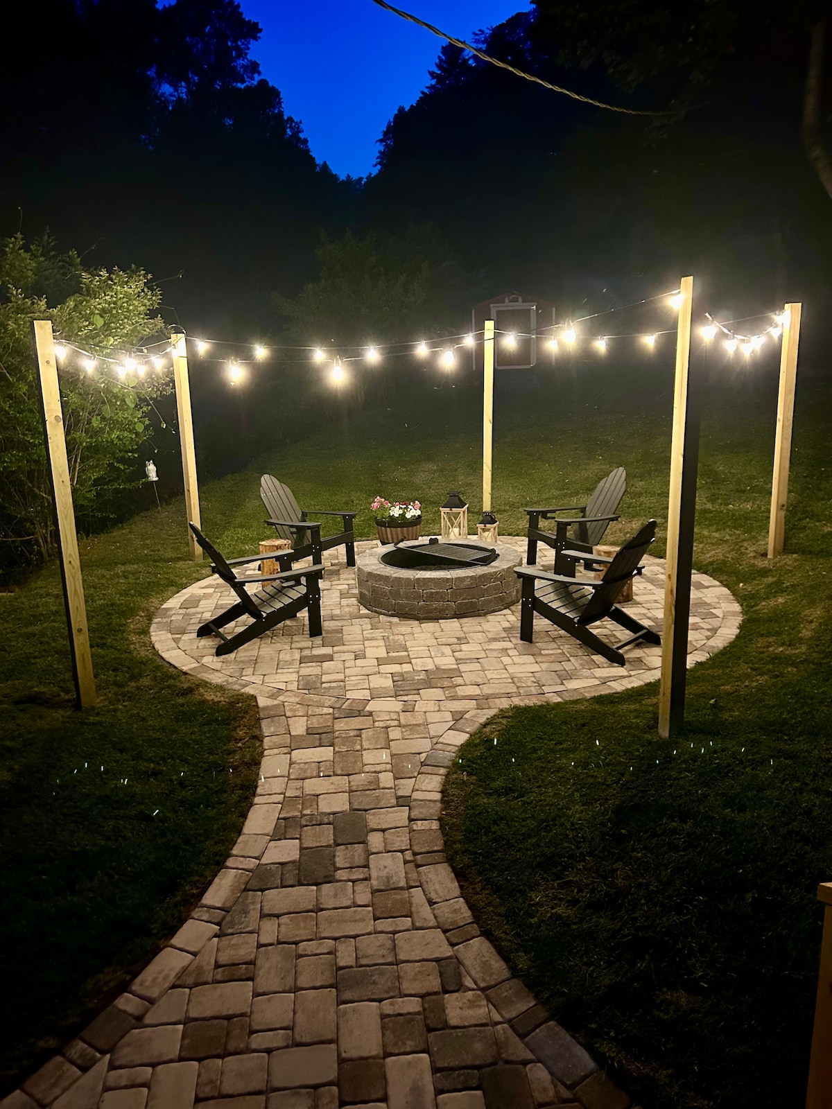 An outdoor fire pit area is illuminated by string lights, creating a serene atmosphere. Four Adirondack chairs surround the circular paved area, which is bordered by well-maintained grass. A winding path leads to the fire pit, enhancing the inviting setting.