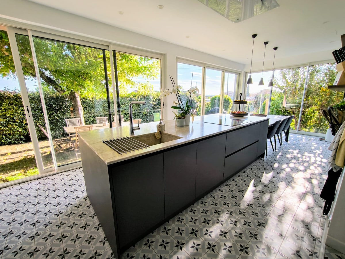 A modern kitchen features a spacious island with a sleek countertop, equipped with a sink and minimalistic fixtures. Large glass doors allow natural light to fill the room while providing views of the outdoor seating area nestled among greenery. A patterned tile floor adds subtle texture to the space.