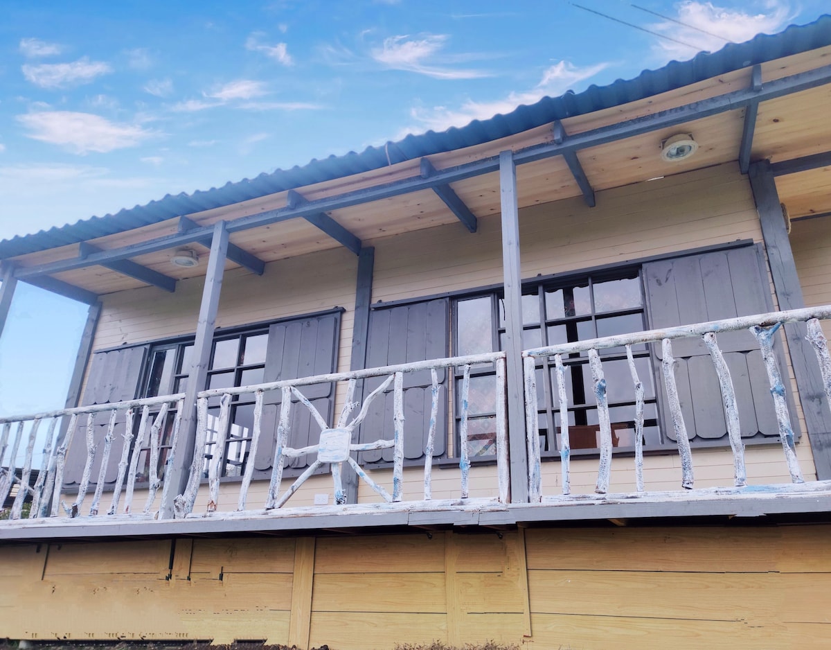 A wooden cabin features a balcony adorned with a railing and several windows, allowing natural light to illuminate the interior. Shutters are painted in a soft hue, complementing the earthy tones of the cabin's exterior under a clear blue sky.