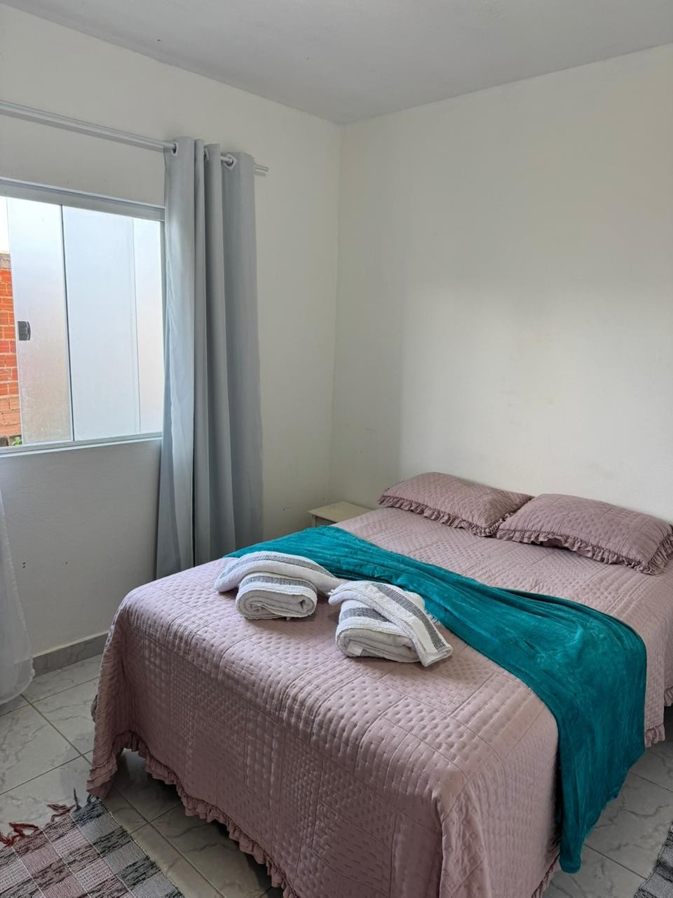 A bedroom features a spacious bed dressed in light pink bedding, complemented by a teal throw. Soft white towels are neatly arranged on the bed. A window with sheer curtains allows natural light to fill the room, enhancing the simple and calm atmosphere.