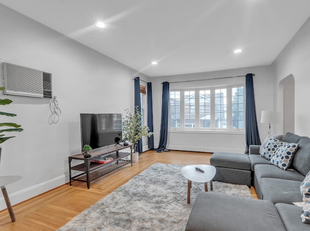The living room displays a comfortable sectional sofa adorned with decorative pillows. A large television is positioned on a sleek media console. Natural light enters through expansive windows, framed by navy curtains, illuminating the light wood flooring and a soft area rug.