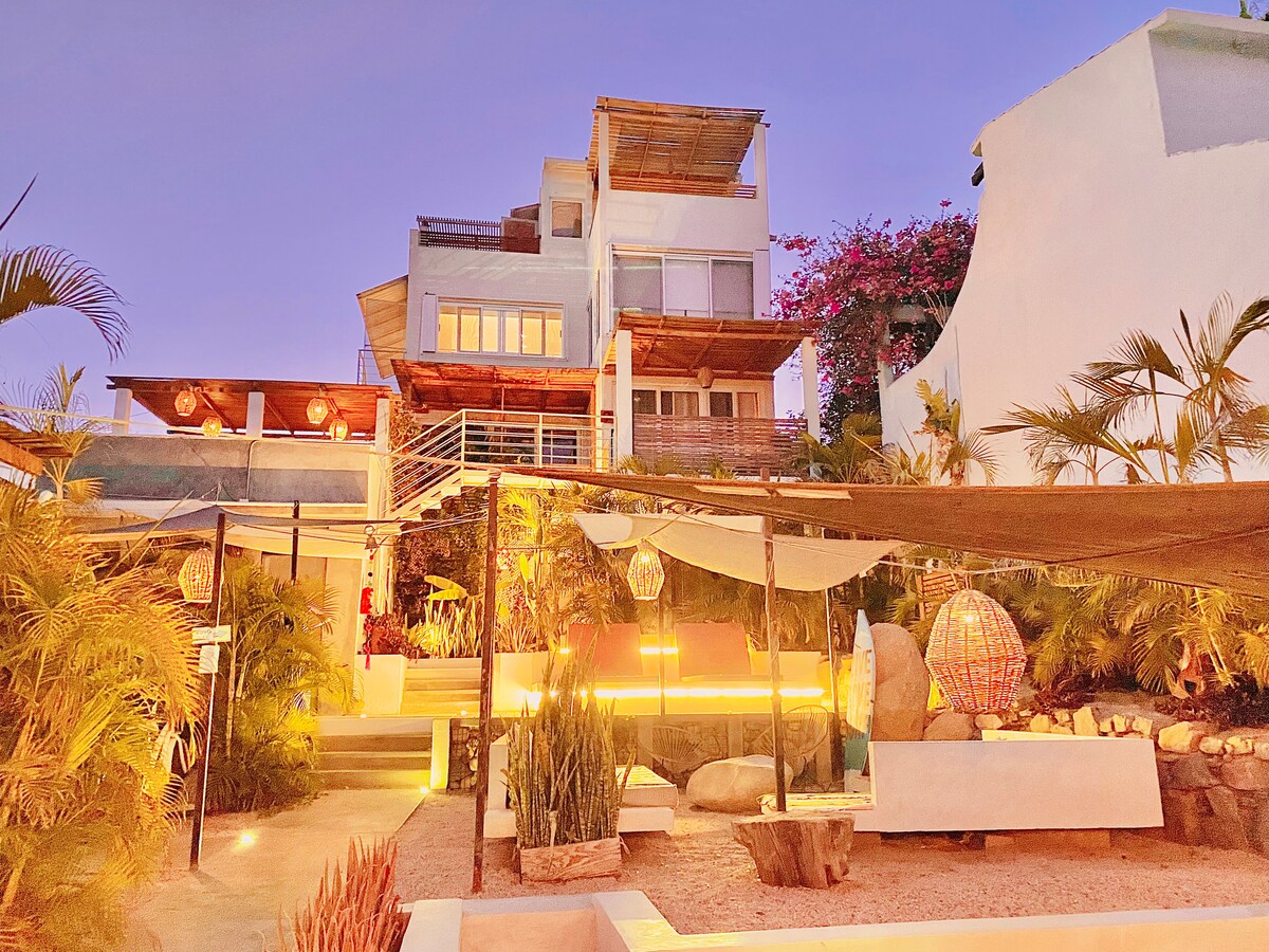 The property is illuminated at dusk, showcasing a multi-level structure surrounded by lush greenery. Natural materials like wood and stone are highlighted, while hanging lanterns and various plant arrangements create a serene outdoor environment.