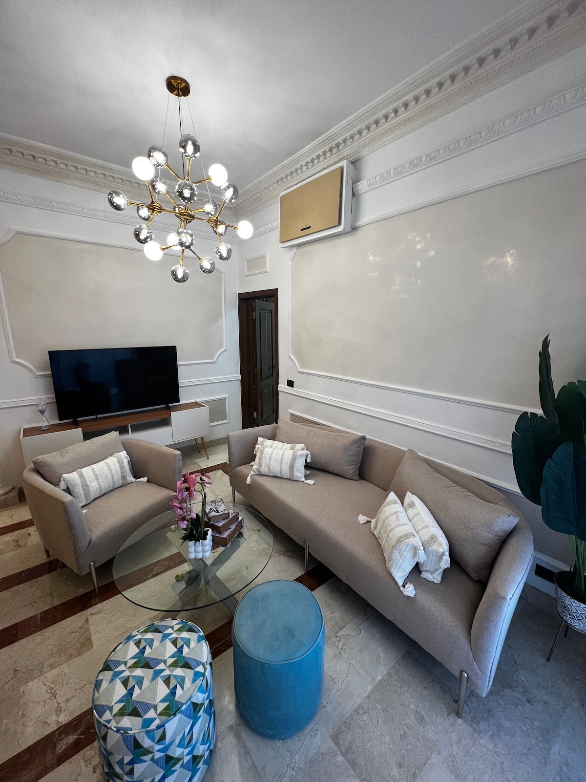 A modern living area features a light-colored sofa and two armchairs, arranged around a circular glass coffee table. A flat-screen television is mounted on the wall opposite, while a decorative plant adds a touch of greenery. Stylish lighting fixtures enhance the overall decor.