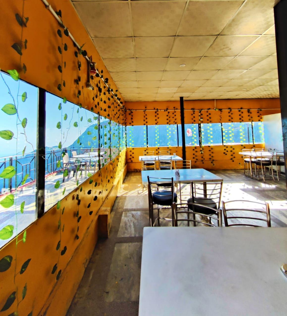 A bright dining area features large windows with leafy decorations, offering sweeping views. Tables with silver metal chairs are arranged throughout, allowing for a spacious dining experience. The walls showcase a warm orange hue, complemented by natural light streaming through the windows.