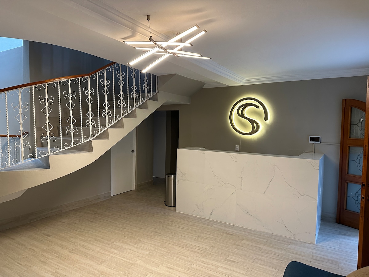 The reception area is characterized by modern design, featuring a marble-fronted desk with a stylized logo. A light fixture with multiple arms adds visual interest to the ceiling. The space is enhanced by a sweeping staircase with a decorative railing, leading to an upper level.
