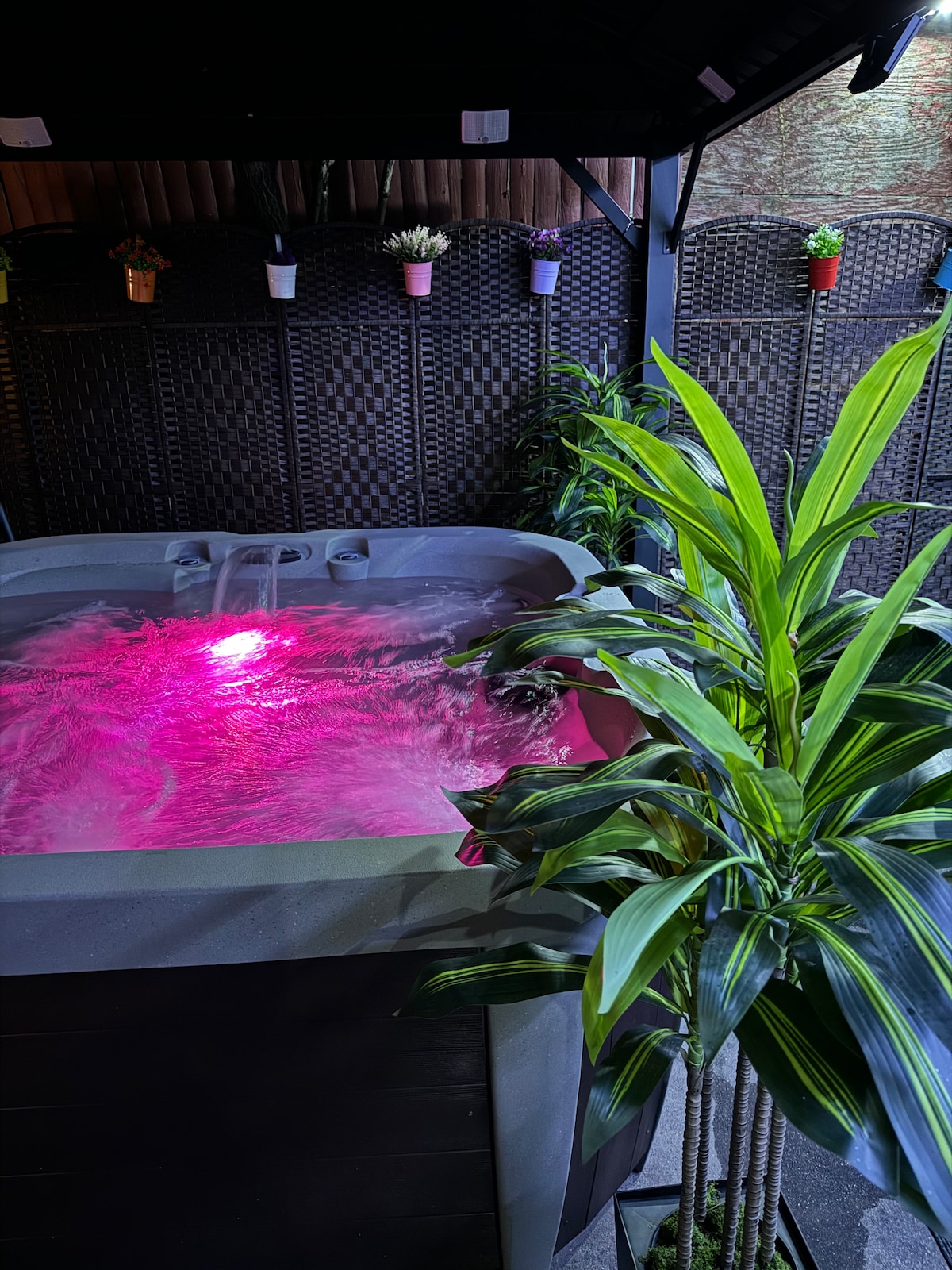 A private hot tub is positioned within a cozy outdoor area, surrounded by lush green plants. Soft pink lighting reflects on the water's surface, creating a serene atmosphere. The background features a textured wall and small potted plants hanging above.