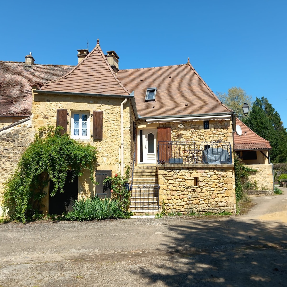 A charming stone house is presented, featuring a sloped roof and warm hues. The entrance is framed by lush greenery, and steps lead to a welcoming door. A small terrace is visible, providing an inviting outdoor space. Clear blue skies offer a tranquil backdrop.