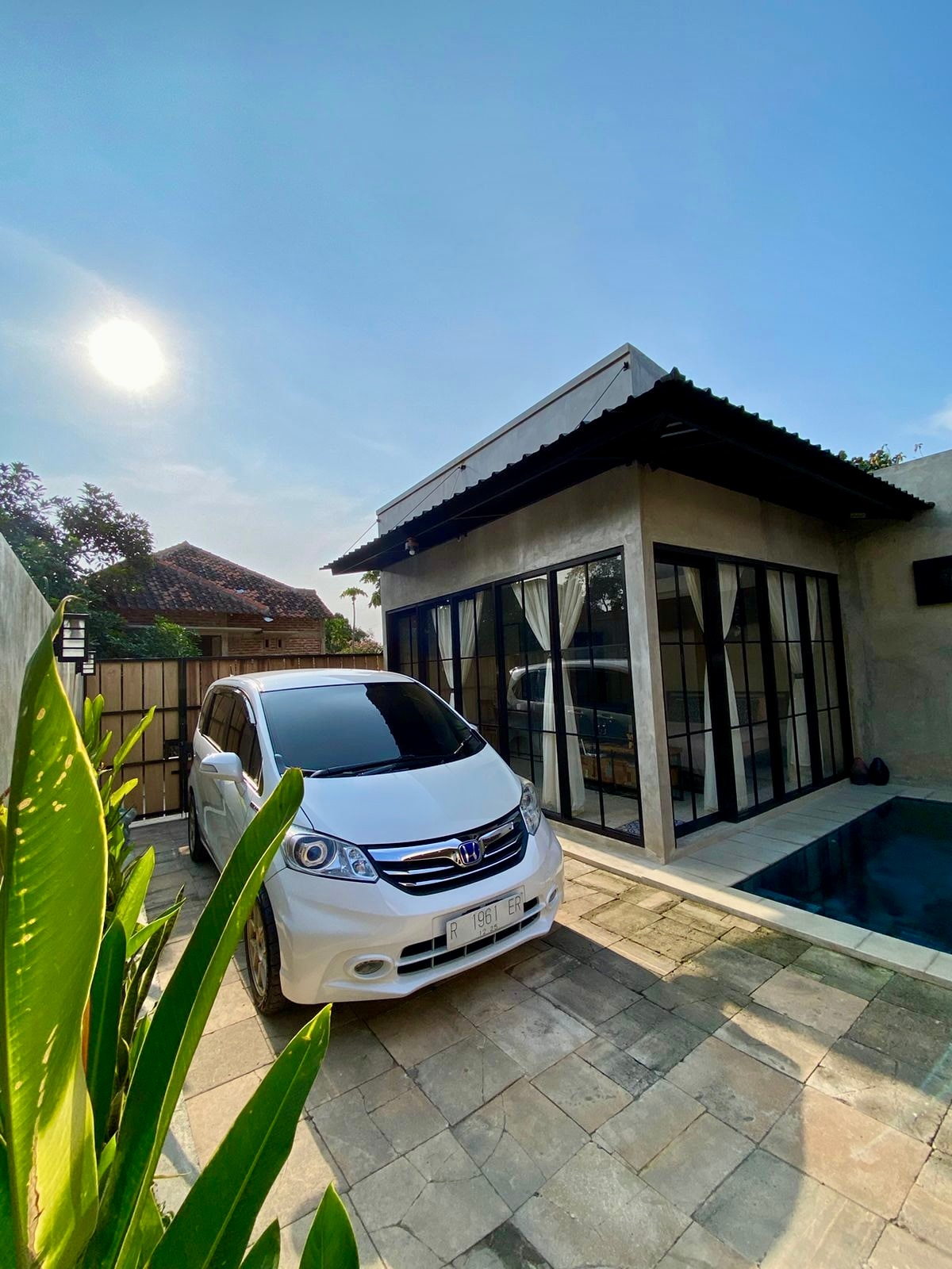 A private house is featured with a pool and a spacious parking area. Large glass windows provide views of the exterior and the surrounding greenery. Sunlight casts a warm glow over the scene, while a white car is parked near the entrance.