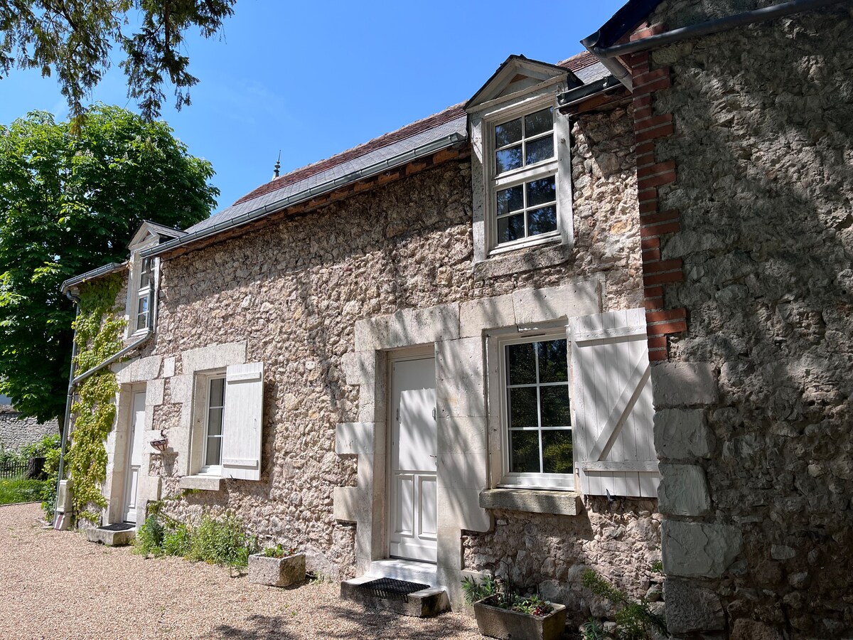 The charming exterior of a stone house is showcased, highlighting its classic architecture with two white doors and several windows. Flanked by lush green trees, the house is set within a peaceful garden, surrounded by gravel pathways and low-maintenance plants.