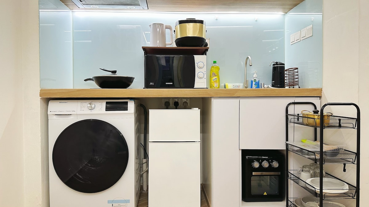 A modern kitchen area is equipped with essential appliances, including a washing machine, refrigerator, microwave, and stove. A light-colored countertop complements the sleek cabinetry. Organized shelves are visible, displaying cooking utensils and cleaning supplies, enhancing functionality and convenience for guests.