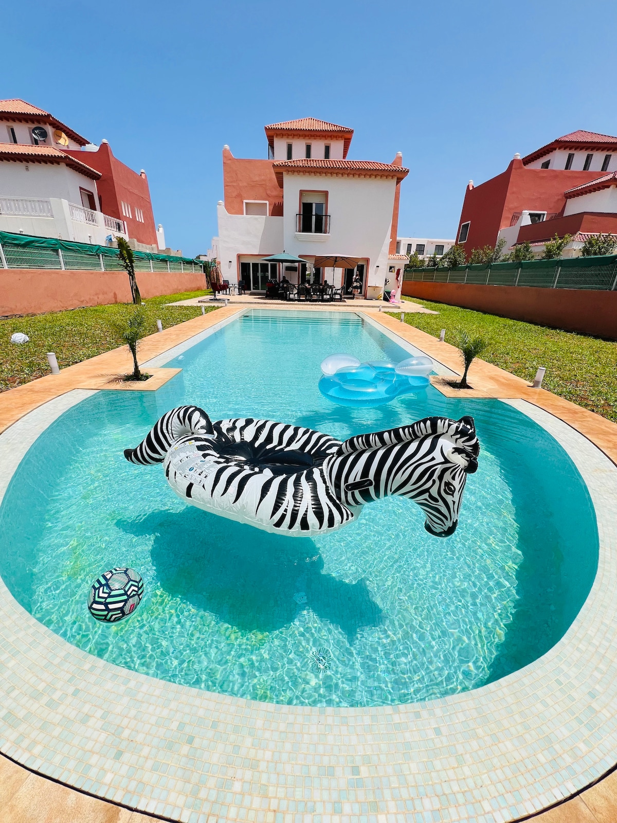 An inviting pool featuring a zebra-striped inflatable floats in the center. Surrounding the pool are well-kept green spaces and palm plants. A two-story villa stands in the background, showcasing its bright exterior against a clear blue sky.