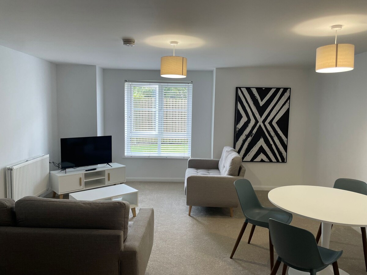 The open living area features a television atop a light-colored media console. Two comfortable gray sofas are arranged around a small coffee table, with a round dining table and chairs positioned nearby. Natural light enters through the window, which overlooks a private outdoor area.