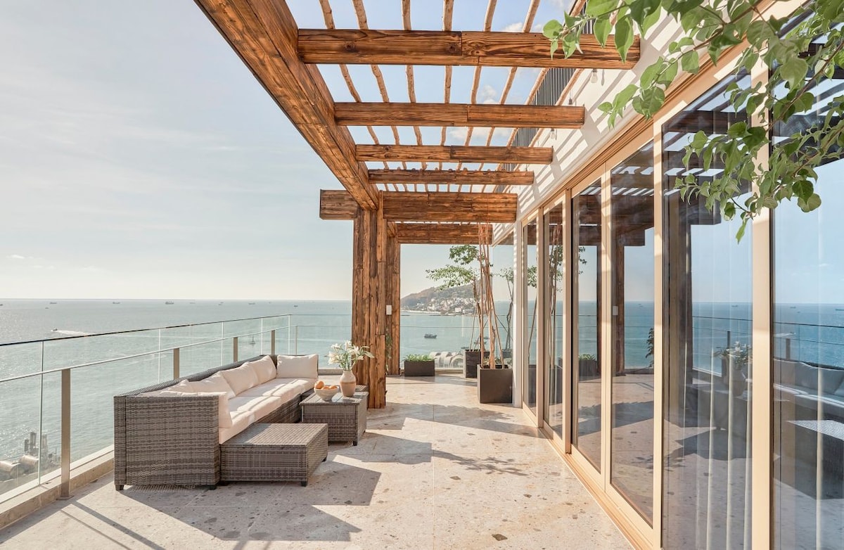 A spacious terrace features a wooden pergola providing shade over a comfortable seating area. The modern outdoor furniture is arranged to face the ocean, with expansive glass doors leading to the indoor space. Views of the sea create a serene backdrop.