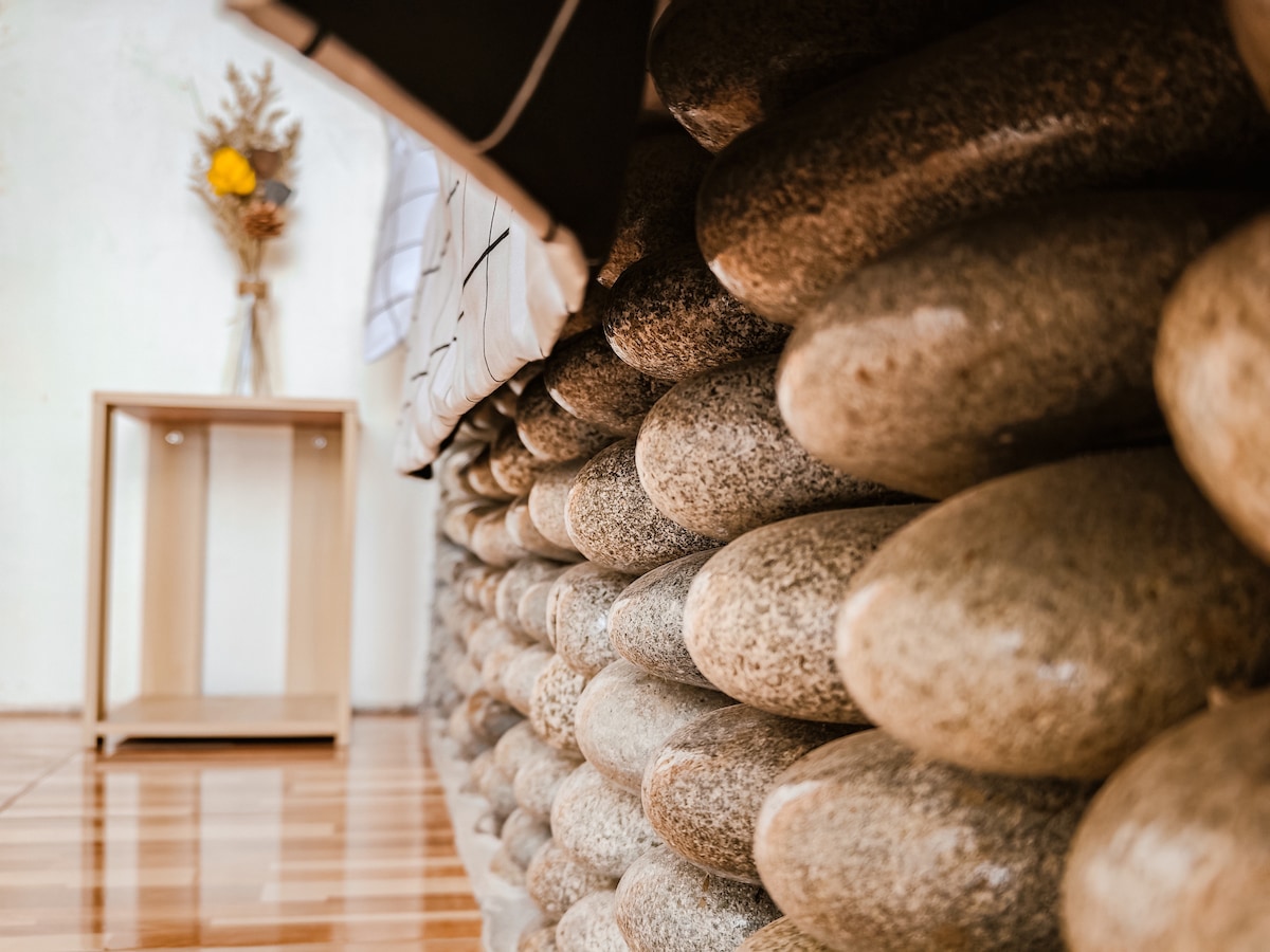 A wall constructed from smooth, rounded stones creates a textured focal point. A wooden side table stands beside it, with a vase holding dried flowers placed atop. Light reflects off the polished wooden floor, enhancing the natural elements present in the space.