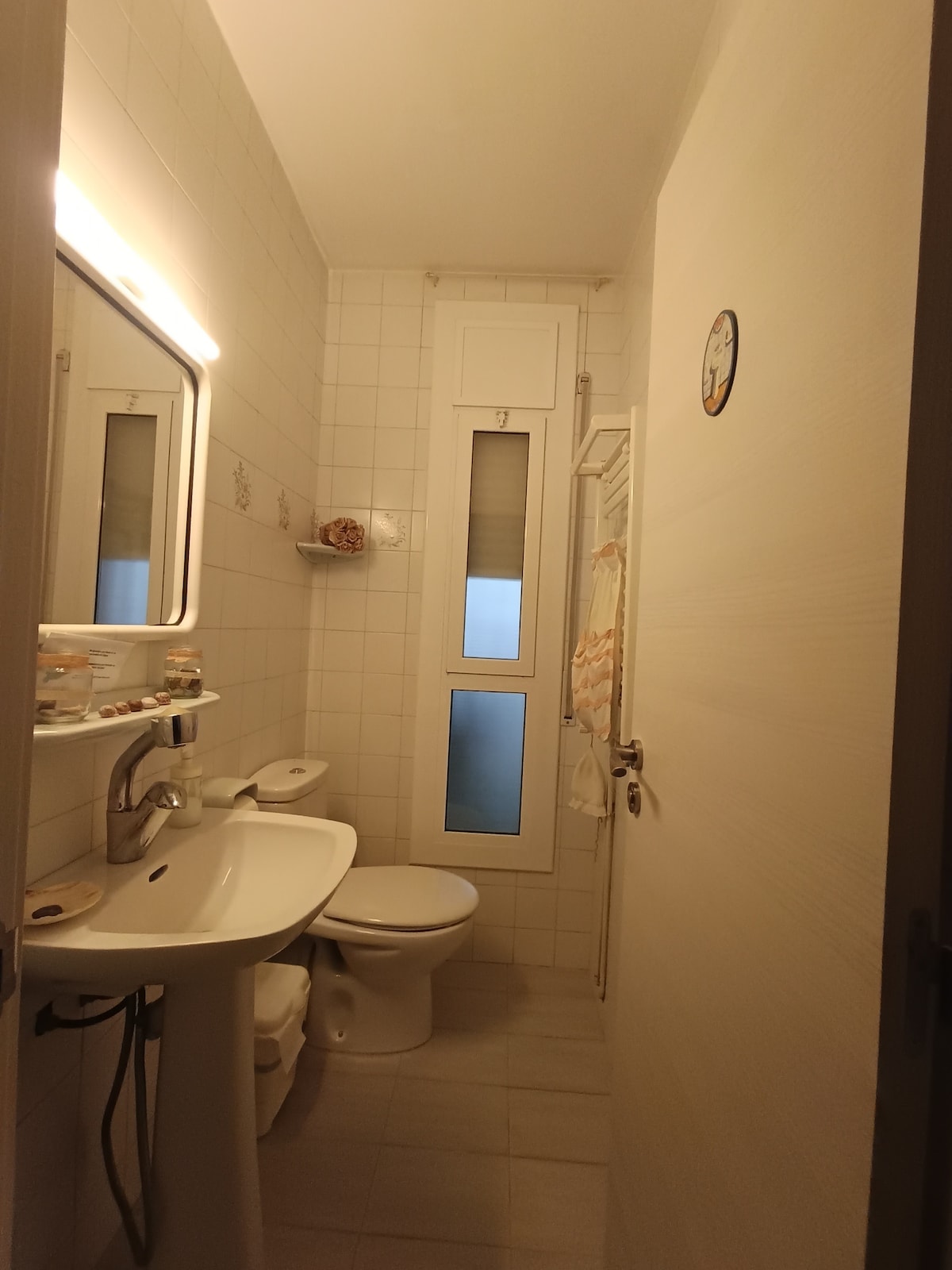 A compact bathroom features white tiled walls and a clean, modern aesthetic. A wall-mounted mirror is illuminated by soft lighting. Essential fixtures include a sink and a toilet, with a narrow window allowing natural light. Towel racks provide convenient storage for linens.