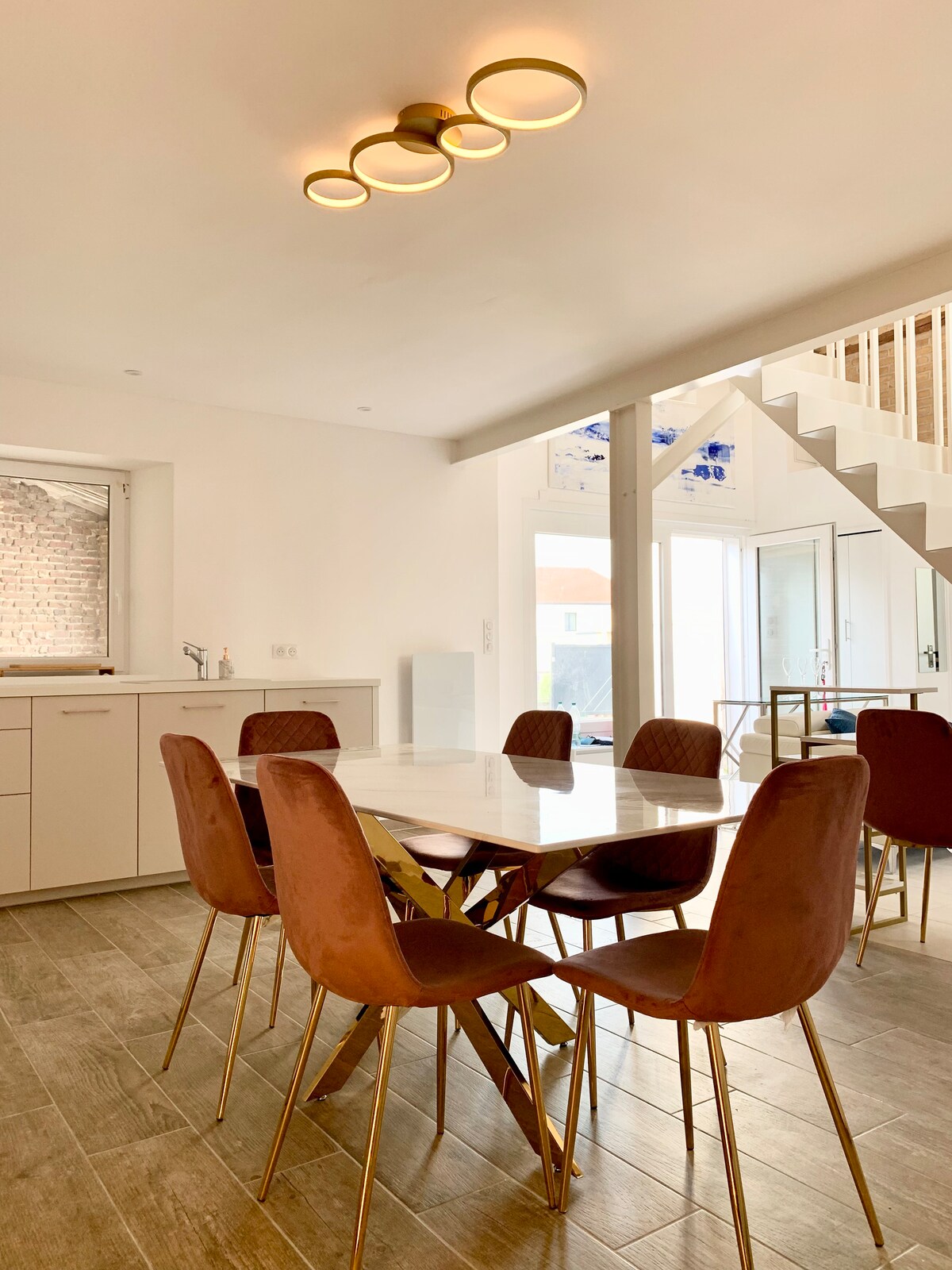 A modern dining area is presented, featuring a sleek white table surrounded by brown upholstered chairs with gold accents. Natural light flows through a nearby window, and a minimalist light fixture hangs from the ceiling, complementing the warm tones of the space.