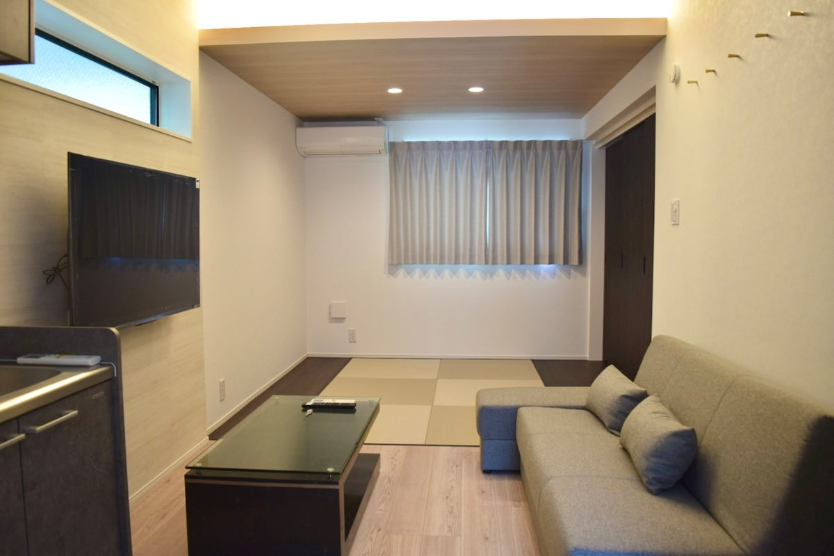 A spacious living area features a gray sofa and a glass coffee table. Natural light enters through large windows adorned with soft curtains. The room's design is complemented by a light wooden ceiling and a carpeted area, with a wall-mounted television visible on one side.