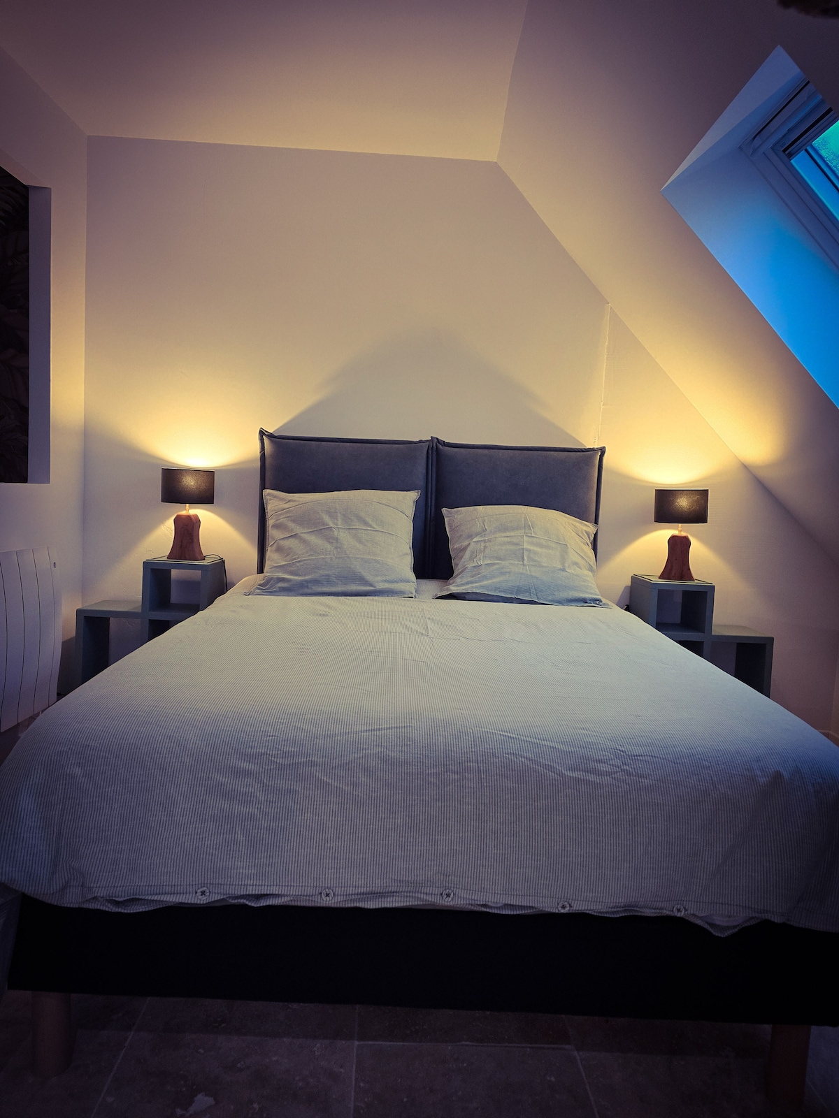 A spacious bed is positioned against a sloped wall, flanked by soft lighting from wall-mounted lamps. Two neutral-colored pillows rest on the inviting linens, creating a calming space. A window is visible above, allowing natural light to enhance the atmosphere.