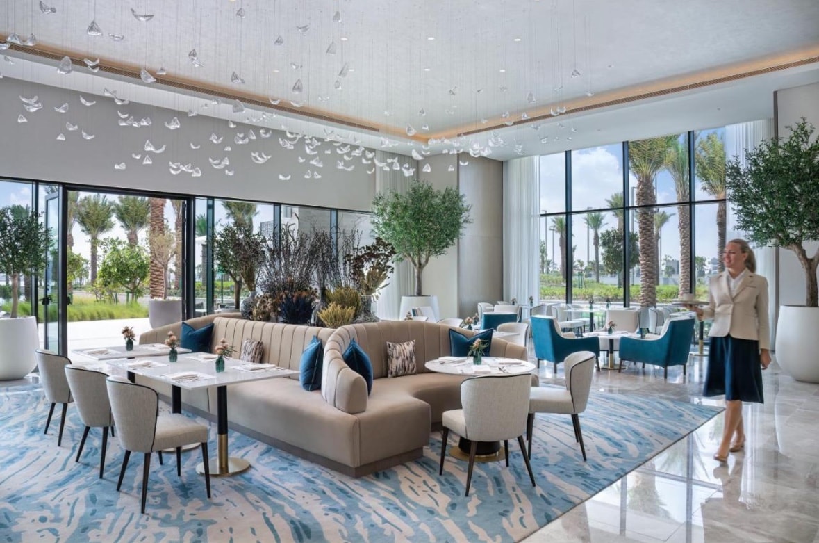 A modern dining area features elegant furnishings, including a soft couch and various dining tables surrounded by upholstered chairs. Large windows provide views of palm trees and greenery outside, while decorative light fixtures hang from the ceiling, creating a welcoming atmosphere.