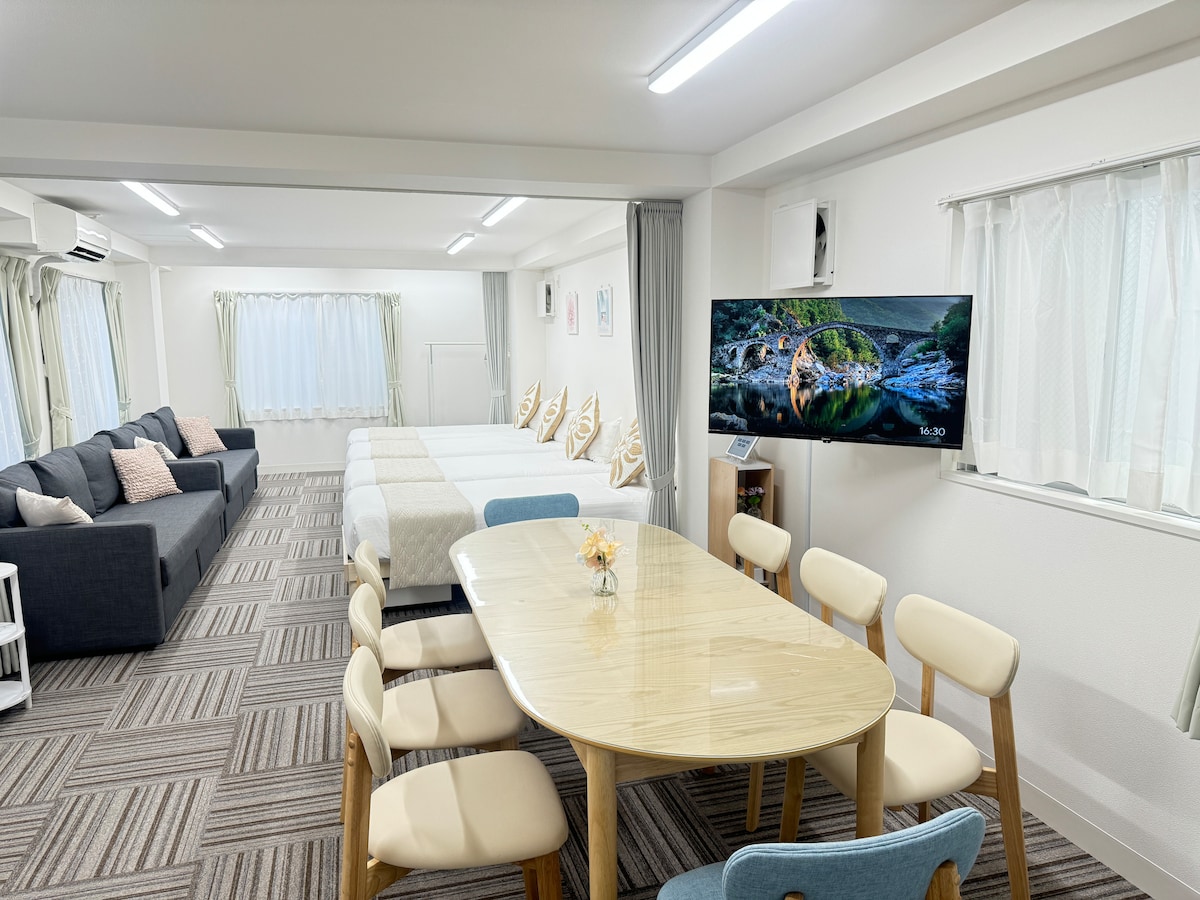 A spacious living area is shown, featuring a large dining table surrounded by eight chairs. Two single beds and a sofa bed are visible, providing ample sleeping options. A TV is mounted on the wall adjacent to a comfortable sitting area, with soft natural light filtering through the windows.