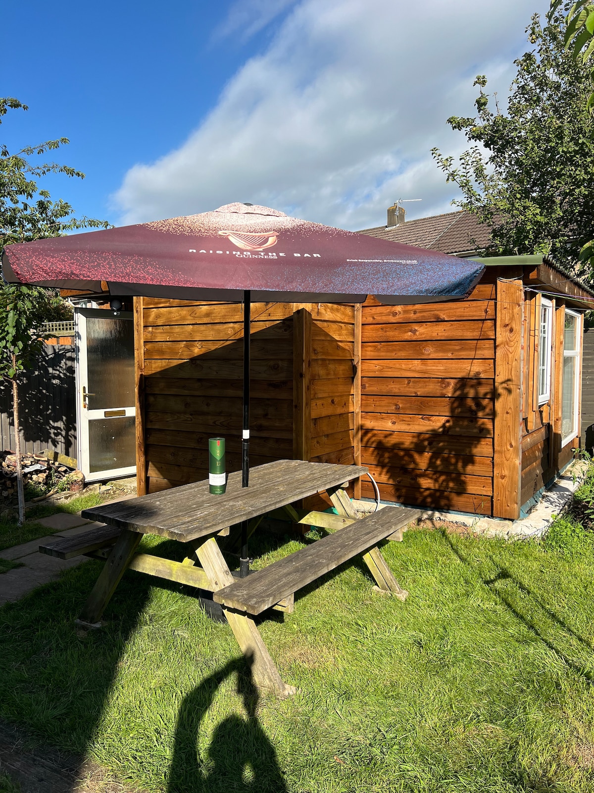 A wooden picnic table with a large umbrella is situated on a grassy area beside a wooden shed. The shed's exterior is composed of natural wood planks, and a patch of blue sky and scattered clouds is visible above.
