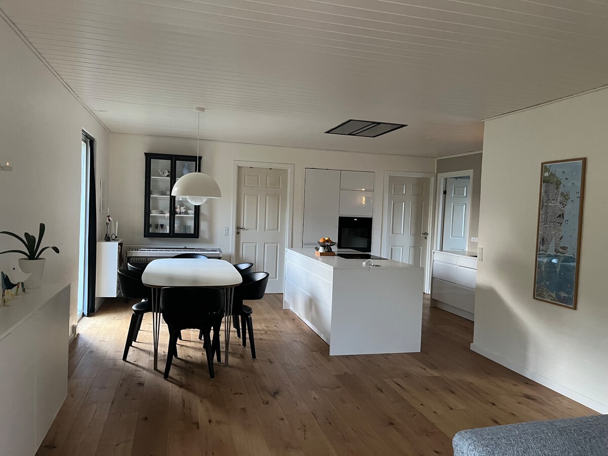 An open-concept kitchen and dining area is presented, featuring a large white table surrounded by black chairs. A stylish pendant light hangs above the table, and a modern kitchen with sleek cabinetry is visible in the background, showcasing a minimalist design.