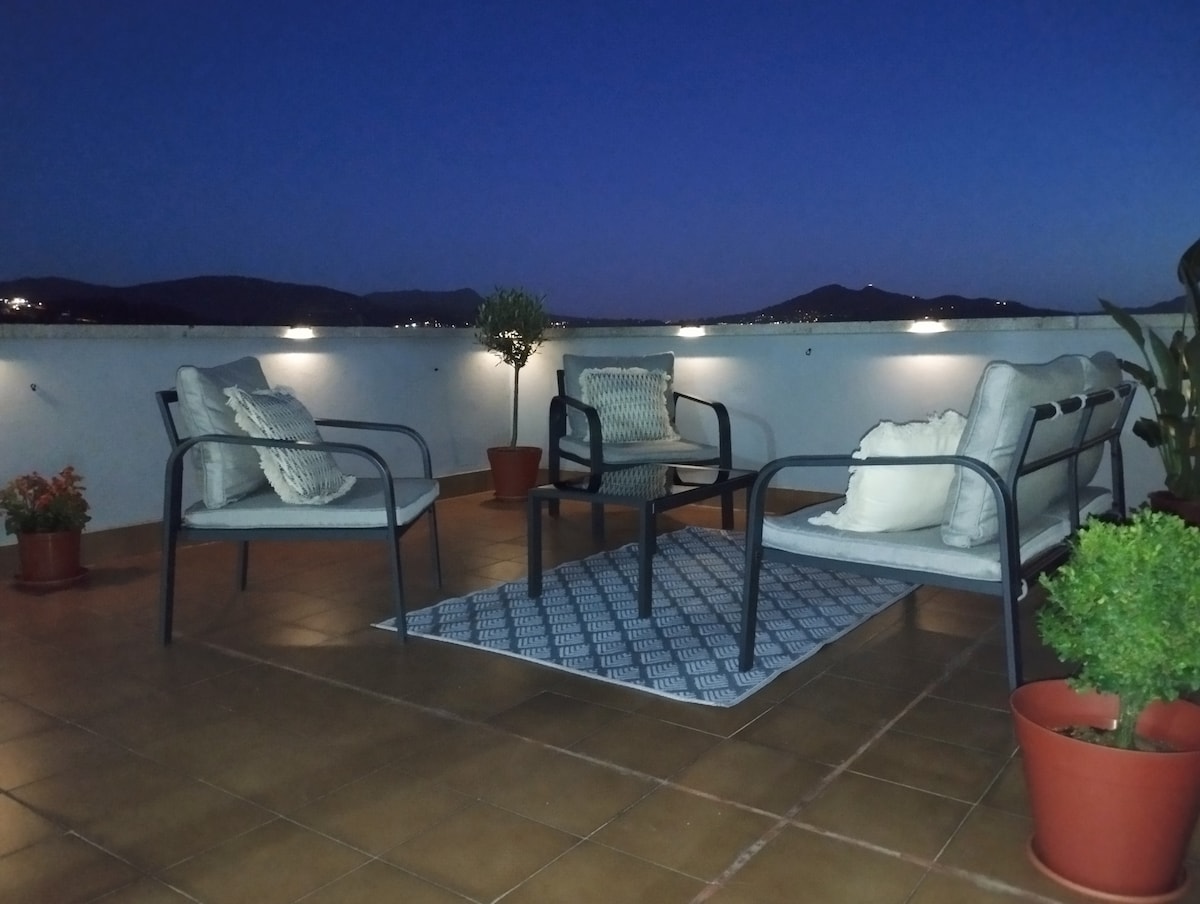 A private terrace is shown at twilight, featuring comfortable seating arrangements with cushions. Potted plants are placed strategically around, adding greenery to the space. A textured rug lies underneath the furniture, complementing the relaxing outdoor atmosphere with views of surrounding hills.
