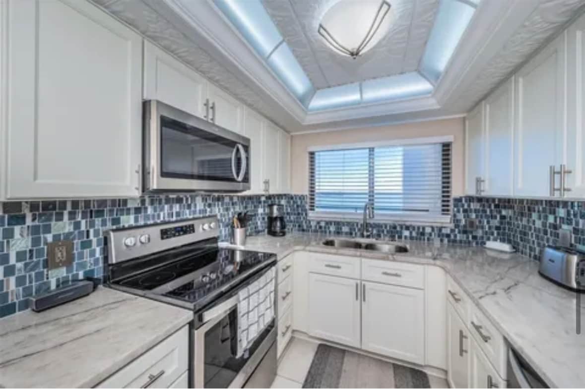 An updated kitchen is presented with white cabinetry and modern stainless-steel appliances. A blend of blue and gray tiles creates a decorative backsplash, while natural light filters in through the blinds-covered window. A marble countertop adds an elegant touch to the space.