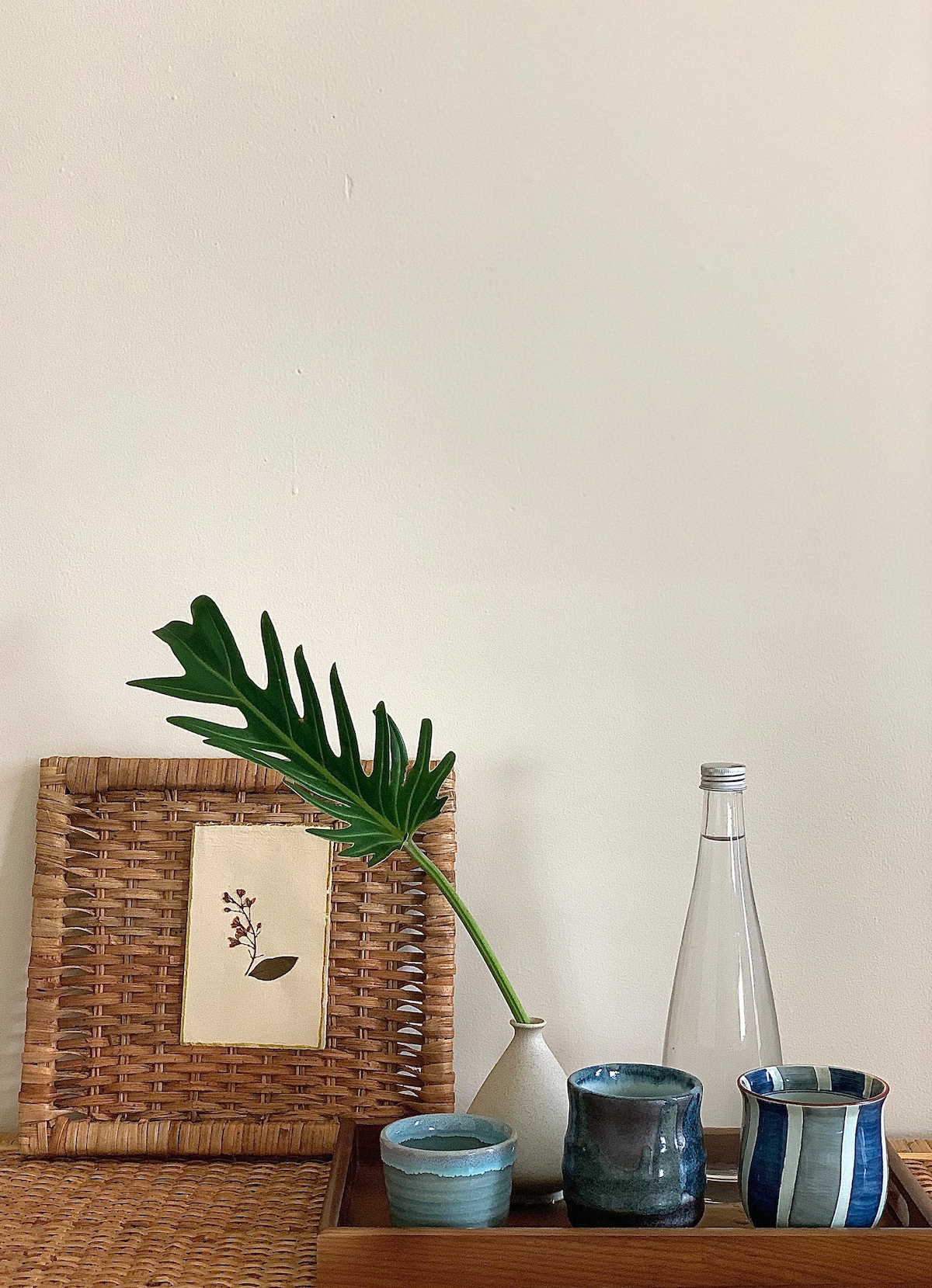 A tray features a selection of handcrafted pottery, including a clear glass bottle and various cups in shades of blue and white. A single green leaf is placed in a small white vase, complemented by a woven wall hanging with a delicate floral design.