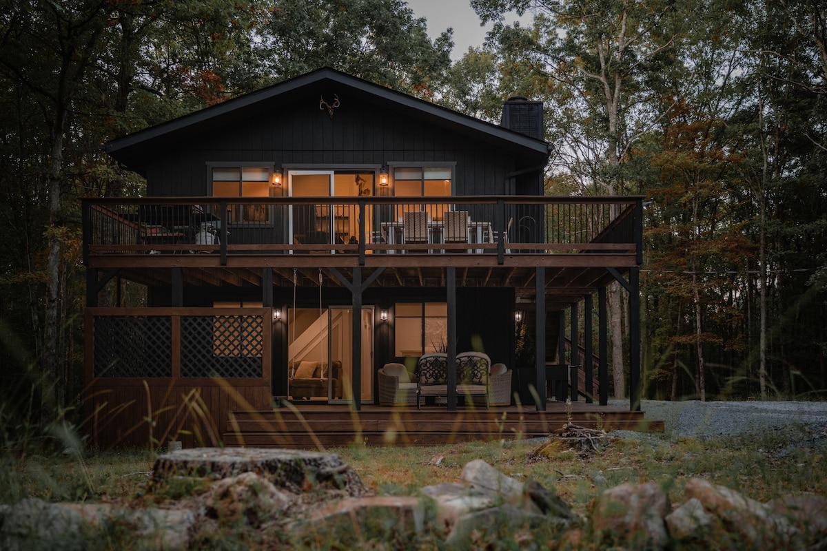 The two-story chalet is surrounded by trees, featuring a large front deck with seating. Outdoor lighting softly illuminates the home, enhancing its welcoming presence amidst the natural setting.