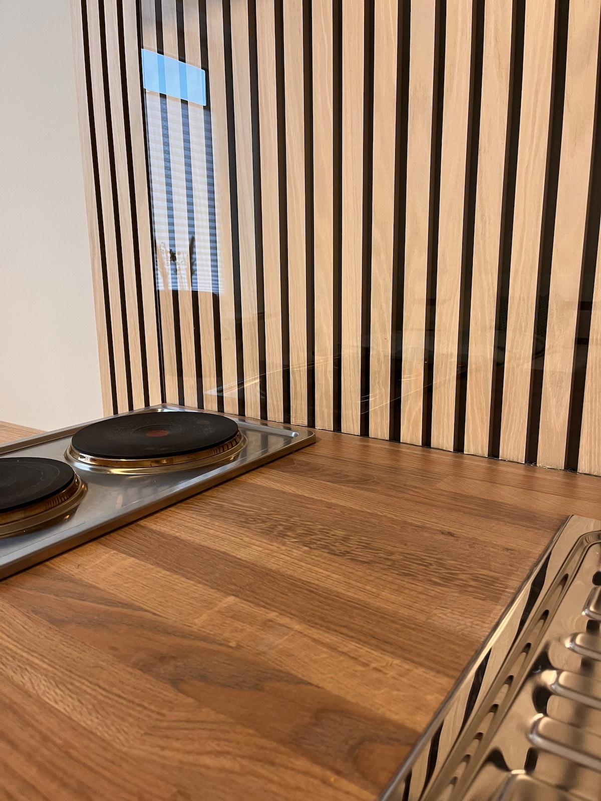 A well-equipped pantry kitchen is showcased, featuring a stainless steel cooktop with two black burners. The wooden countertop exhibits a natural finish, complemented by vertical wooden paneling in varying tones. A glimpse of light is visible through a nearby window.