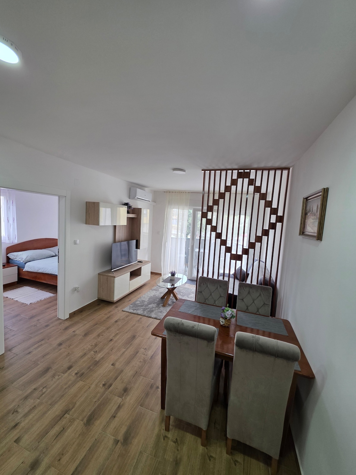 A modern living space is presented, featuring a dining area with a wooden table and upholstered chairs. A partition with a geometric design separates the dining area from a cozy bedroom, where a spacious bed is visible. Natural light enhances the inviting atmosphere.