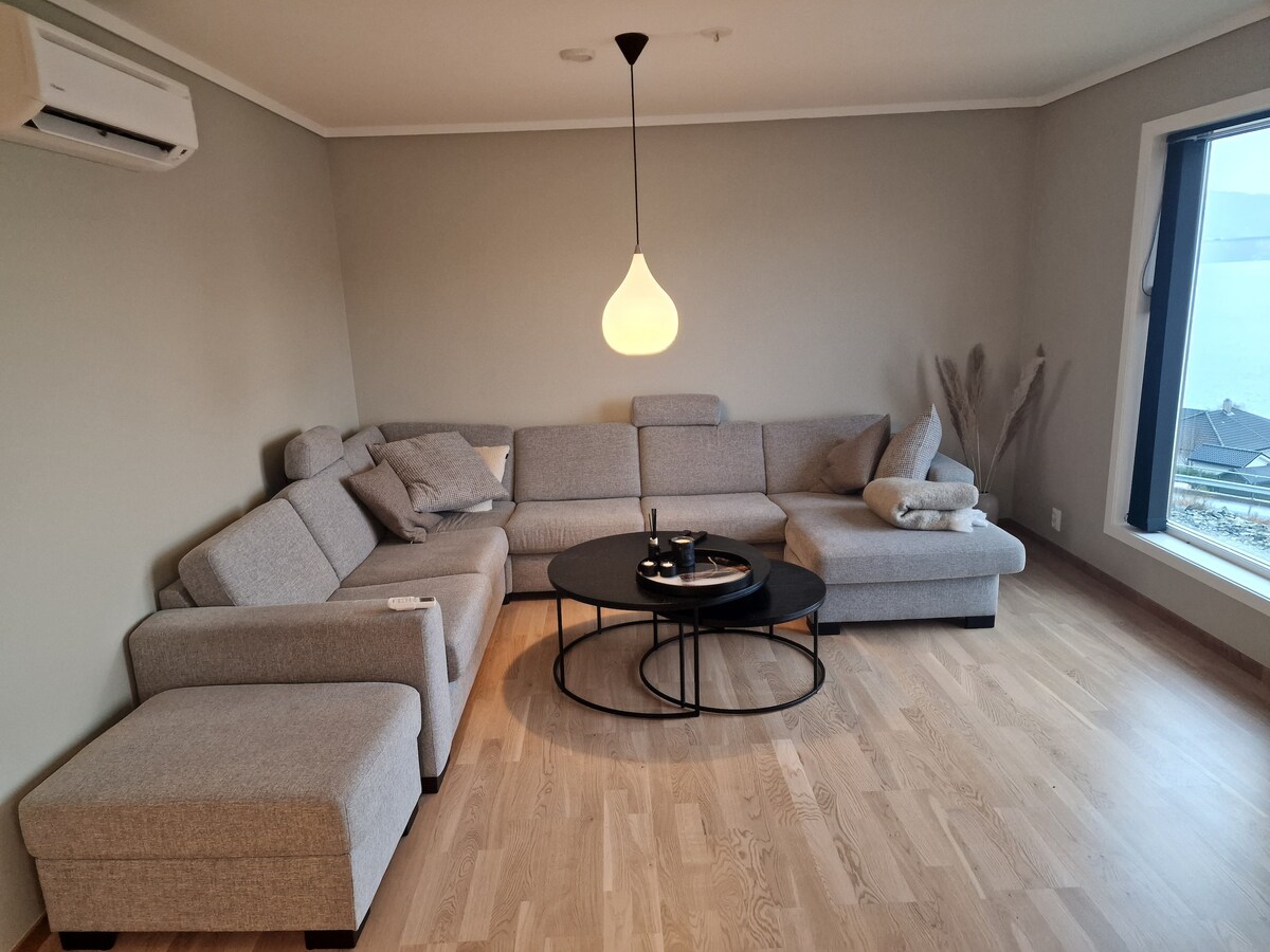 A modern living area features a spacious grey sectional sofa adorned with multiple throw pillows. A round black coffee table sits in front, attended by decorative items. Natural light fills the space through a large window, complemented by minimalist decor.