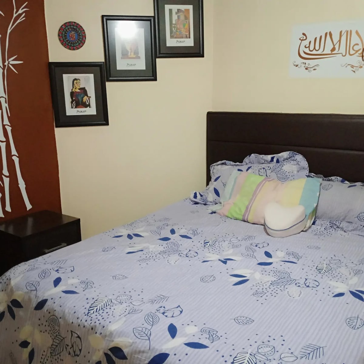 A comfortable sleeping area is furnished with a spacious bed adorned with patterned bedding featuring blue and white floral designs. Framed artwork is displayed on the wall, complemented by a decorative wall feature. Soft lighting illuminates the warm-toned room.