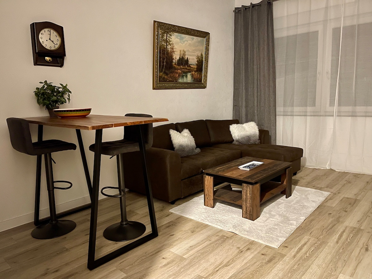 The living area features a brown sectional sofa complemented by two decorative pillows and a coffee table crafted from wood. A dining table with two stools is present, and a wall clock adds a classic touch. Natural light filters through sheer curtains.