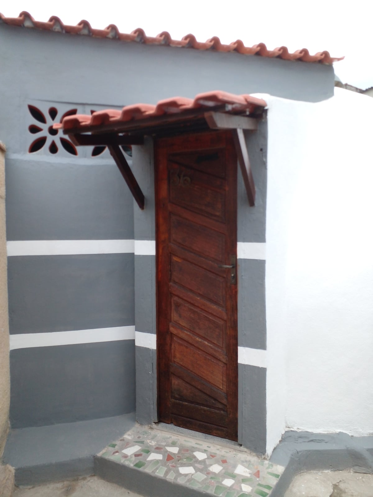 A wooden door with a diagonal pattern is framed by a gray and white exterior wall. A decorative vent is visible above the door, while a small overhang with exposed beams provides shelter. The floor features a simple tile pattern, adding subtle character.