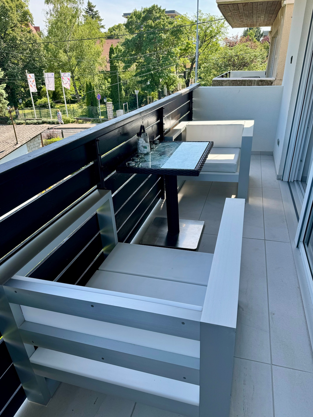 A private terrace is shown, featuring a glass-topped table and two light-colored armchairs. The setting is complemented by a view of greenery in the background, while the sleek railing provides a modern touch to the space.