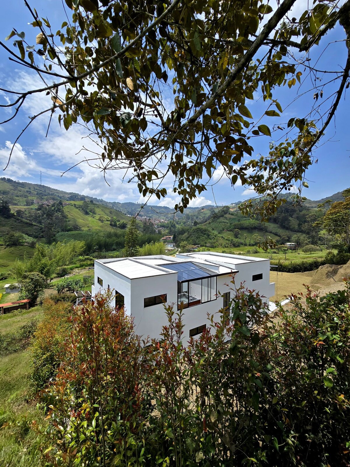 A modern white house is framed by lush greenery and trees, set against a backdrop of rolling hills and a clear blue sky. Natural light reflects off the building's large windows, highlighting its clean lines and contemporary design.