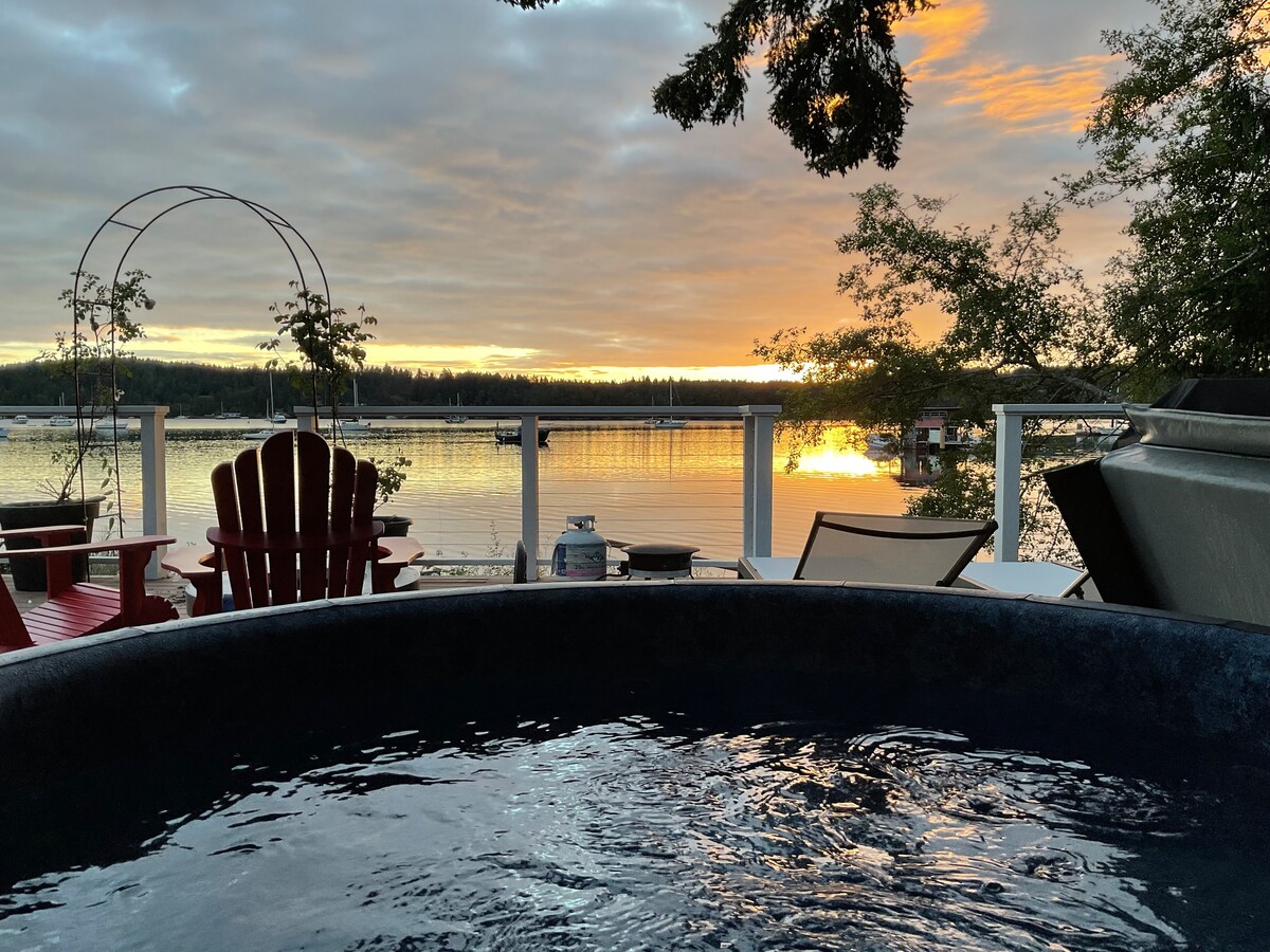 A jacuzzi is positioned at the edge of a deck overlooking serene waters. Vibrant orange and yellow hues of the sunset are reflected on the water's surface. Red adirondack chairs are visible in the background, enhancing the tranquil outdoor setting.