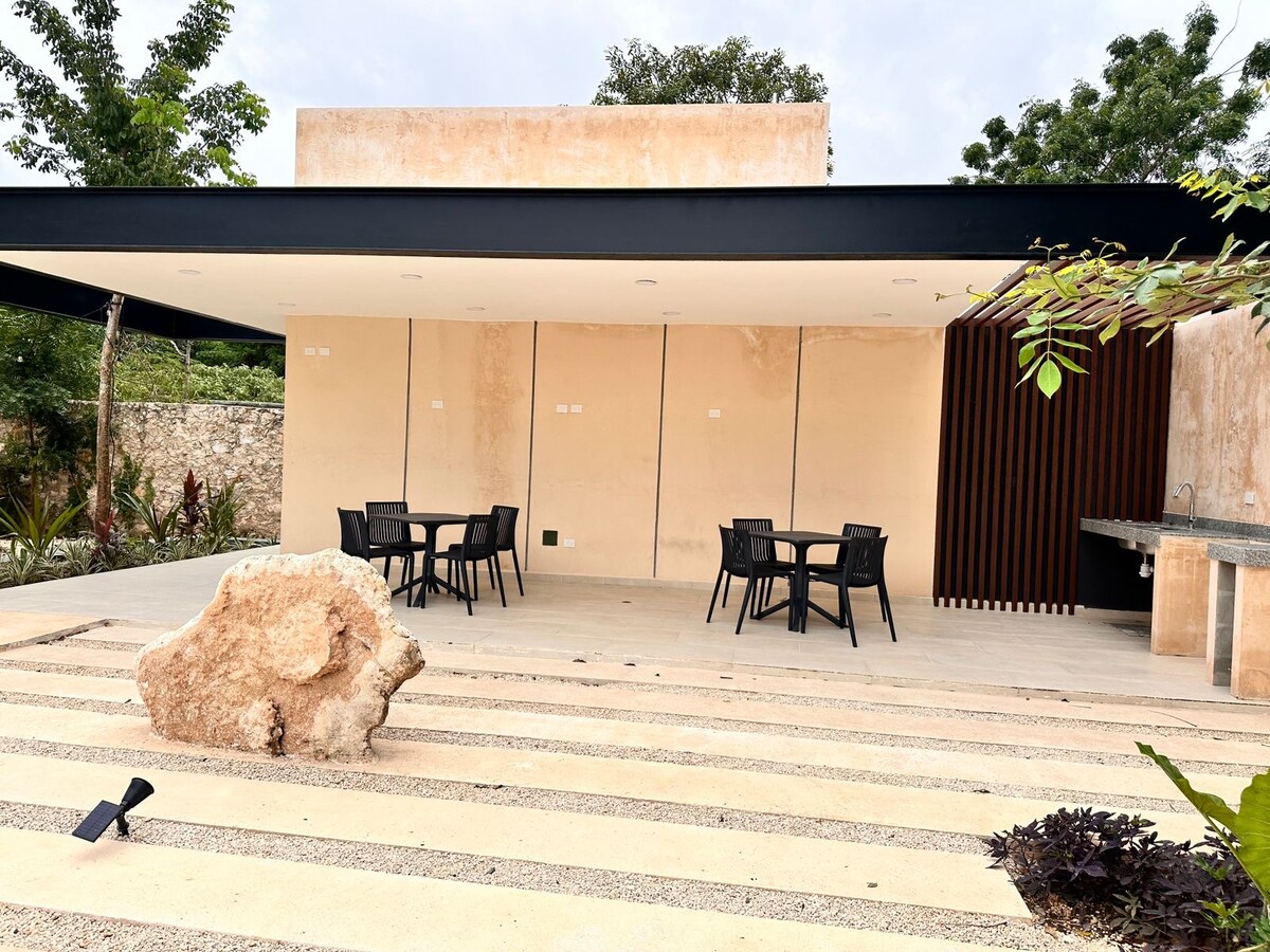 An outdoor patio area is displayed, featuring a wooden deck with a seating arrangement that includes several black chairs and a table. Natural stone elements are present, along with a backdrop of lush greenery and a textured wall, providing a serene environment.