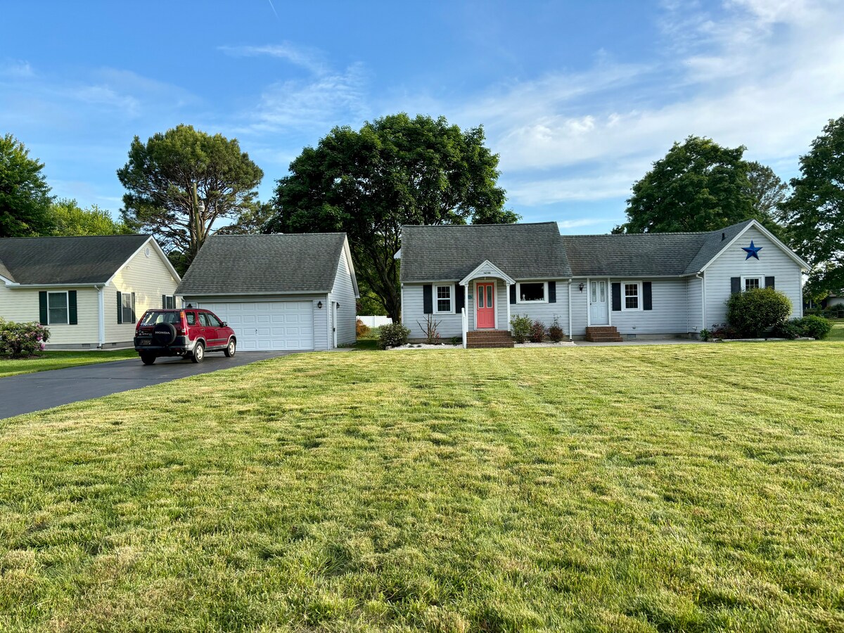 The exterior of a charming single-story bungalow is showcased, featuring a bright pink door and a star accent above it. A well-maintained lawn stretches across the front yard, bordered by trees, with a paved driveway and a garage visible on the left.