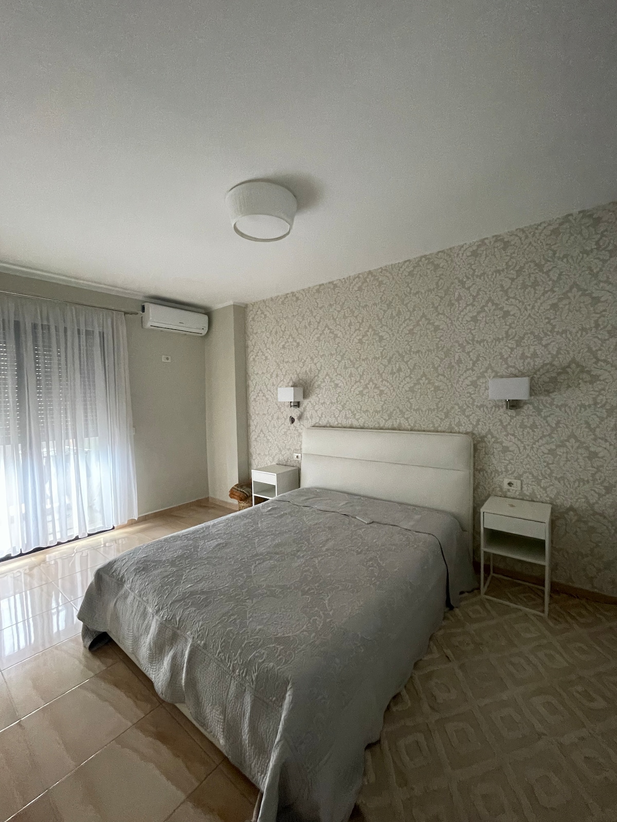 A light and airy bedroom features a spacious bed with a soft grey coverlet. Two bedside tables with lamps are positioned on either side of the bed. Natural light enters through a large window, dressed with sheer curtains, illuminating the patterned wallpaper and sleek flooring.