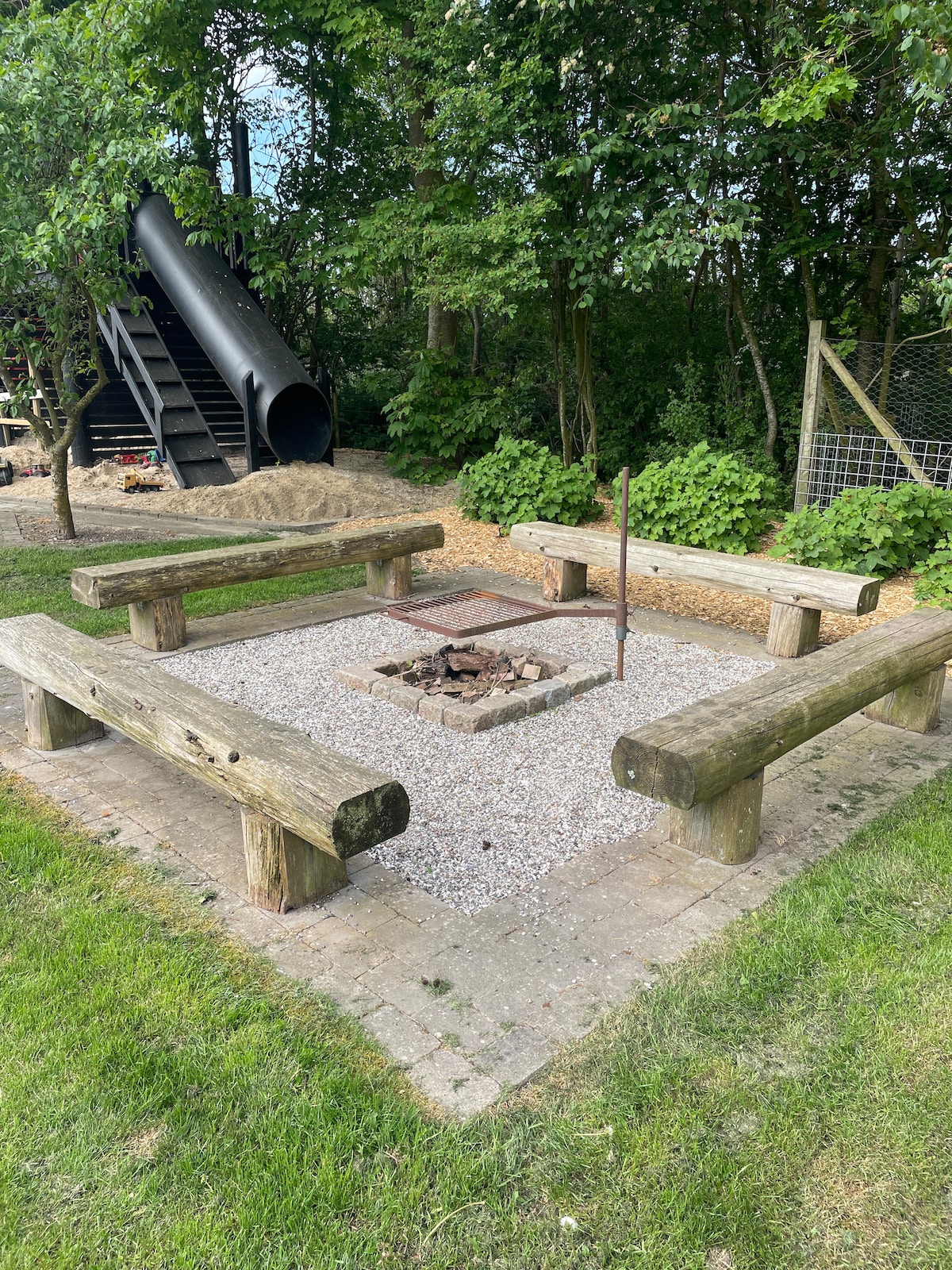 An outdoor fire pit area is surrounded by four log benches, offering a communal space for relaxation. The ground is paved with gravel, and a small fire ring is positioned at the center. Lush greenery and a slide are visible in the background.