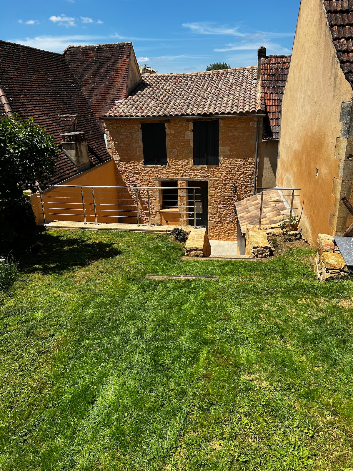 A well-maintained outdoor space is featured, showcasing a neatly trimmed lawn leading up to a charming stone house. The house features large windows with shutters, and a small path is visible, bordered by a low stone wall and surrounded by greenery.