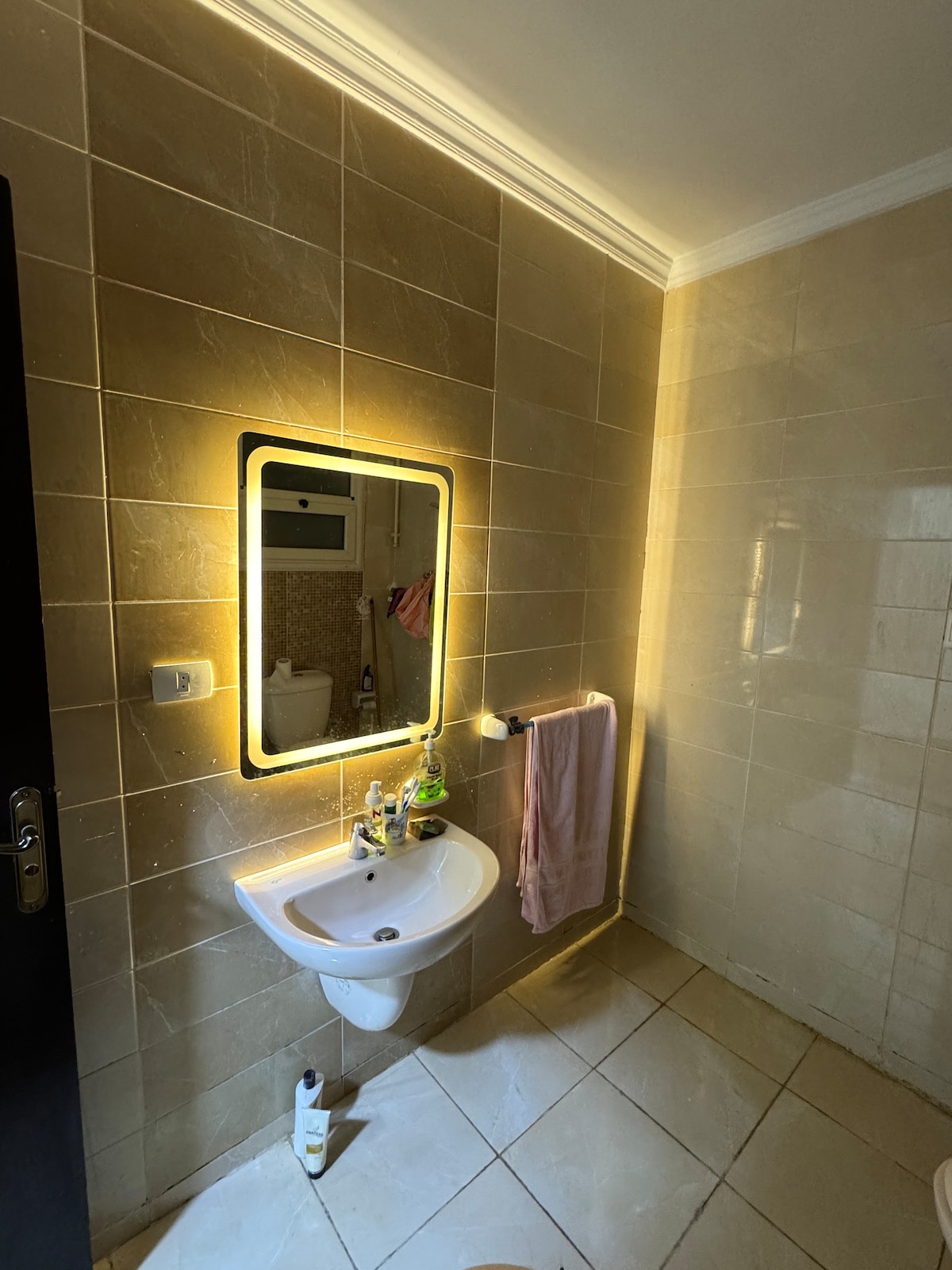 A modern bathroom is presented, featuring a well-lit mirror with a glowing edge above a sleek sink. A towel hangs on a nearby rack, and essential toiletries are neatly arranged on the counter. Neutral tiled walls and floor create a clean, spacious appearance.