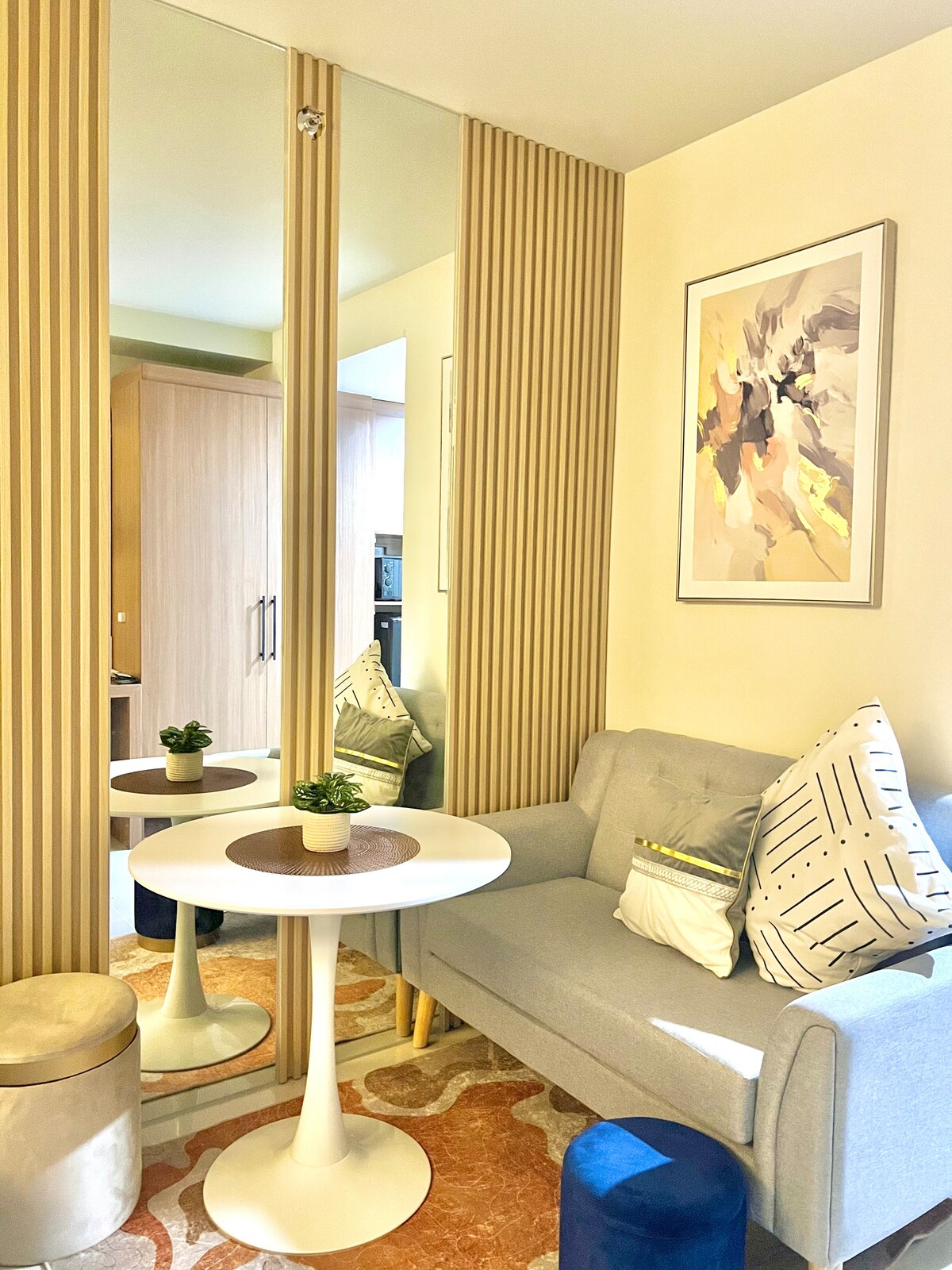 A compact seating area features a light gray sofa paired with colorful cushions. A round white table rests on a patterned rug, with a small potted plant at its center. Two mirrors reflect the space, enhancing the bright, airy atmosphere.